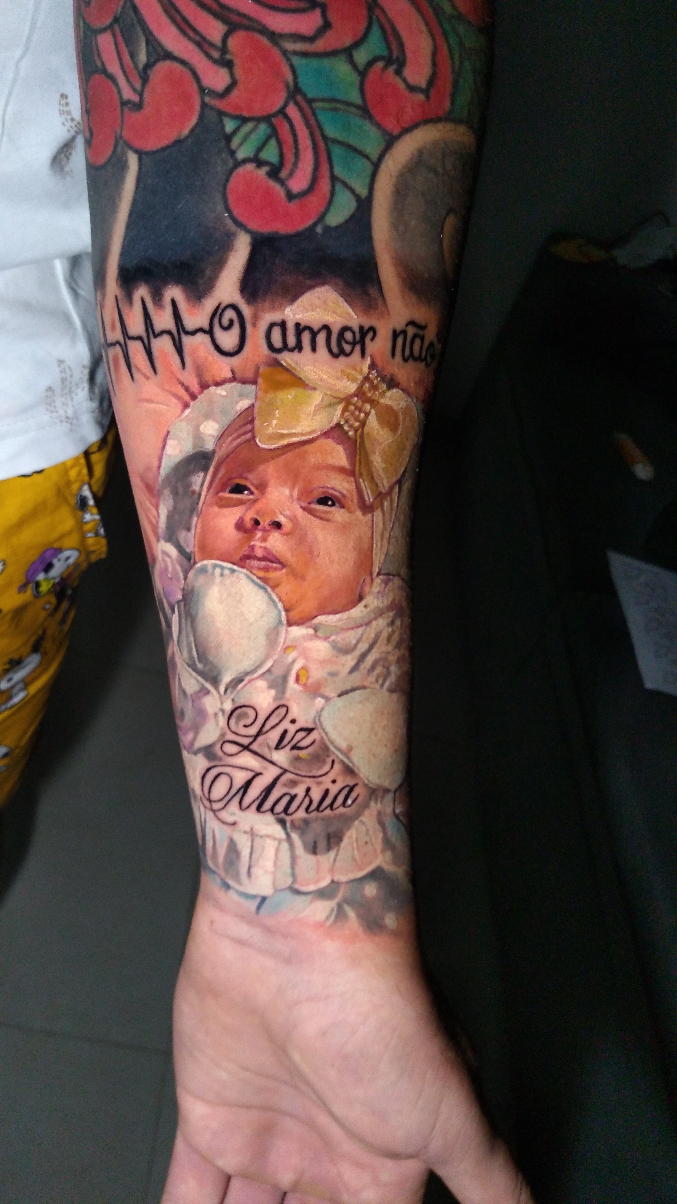 Retrato realista colorido de bebê Liz Maria tatuado no antebraço com nome em lettering e linha de batimento cardíaco — foto com filtro polarizado